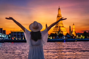 A happy tourist woman enjoys the view to the illuminated Wat Arun temple in Bangkok, Thailand,...