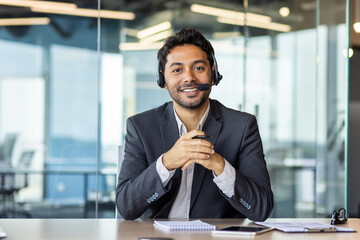 Close-up portrait of a young Indian man sitting in the office at the desk wearing a headset, smiling and talking to the camera.