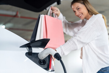 Young woman travel with EV electric car to shopping center parking lot charging in downtown city...