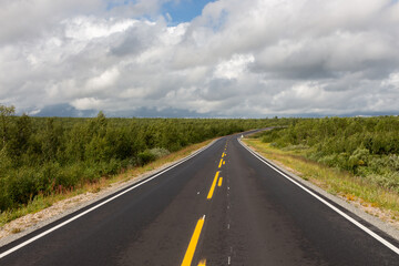 Newly paved road with endless yellow stripes between forests and blue sky with big clouds