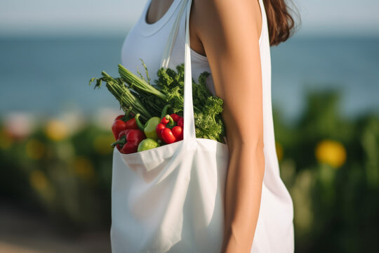 Close up view of young woman wearing T-shirts mockup holding a reusable white tote bag with colorful vegetables in background of local farmer market. Shopping and travel lifestyle concept.