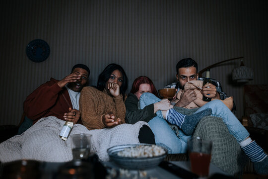 Multiracial friends watching scary movie together while sitting on sofa in cabin