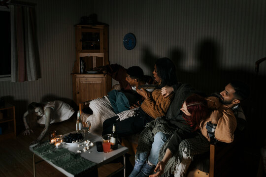 Frightened multiracial friends looking at spooky ghost while watching scary movie at cabin