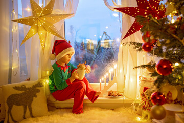Toddler child, cute blond boy, sitting on the window in pajama, looking out for Satna Claus,...