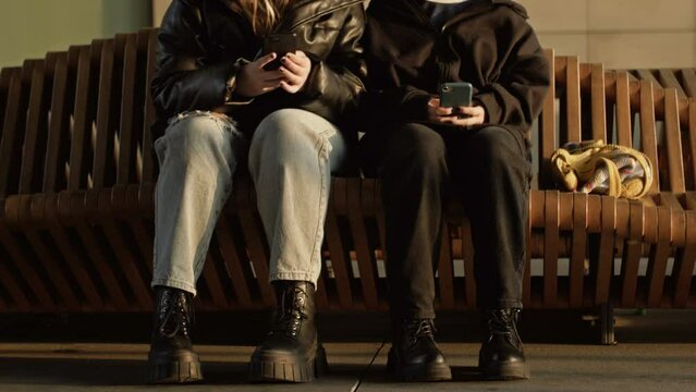 Two girls are sitting on a bench at the train station with smartphones in their hands.