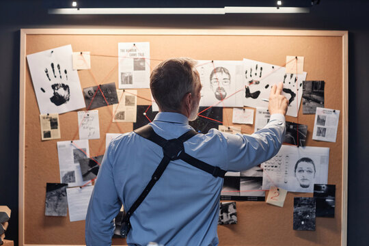 Back view of senior detective standing by evidence board and studying leads in investigation