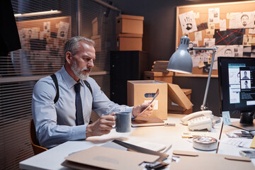 Portrait of handsome male detective studying evidence and drinking coffee at workplace in office,...