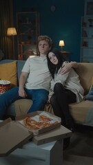 Portrait of teenage couple spending leisure time. Boy and girl sitting on the sofa eating popcorn and watching tv and hugging. Vertical photo.