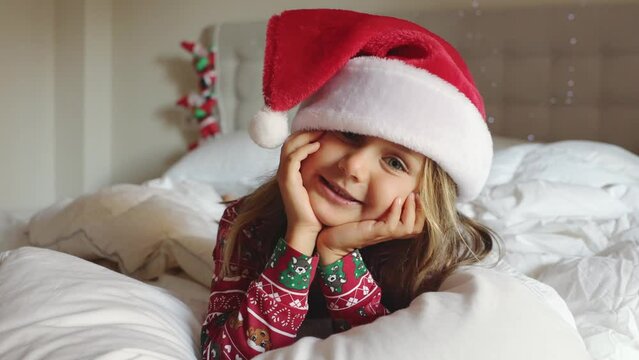 Merry Christmas Holidays 2023 New Year. Happy smiling child girl lies in cozy bed under a white fluffy blanket decorated home lights. Toddler in red Santa hat with pompon and pajamas. Good morning