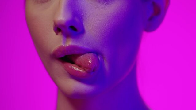 Zmysłowe zbliżenie kobiecych ust z językiem wystawionym na bok w kolorowym świetle fioletowo-niebieskim na r&oacute;żowym tle &mdash; slow motion 4K, sensualność, detal i kobiecy styl