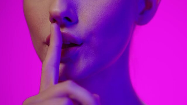 Zbliżenie kobiecych ust z palcem przyłożonym do warg w geście ciszy na r&oacute;żowym tle z niebieskim światłem slow motion 4K, tajemnica, gest i kobieca ekspresja