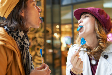 Happy women joking with blue tongue lollipops with urban lights background - obrazy, fototapety, plakaty