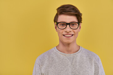 joyful young man in white attire with glasses looking at camera smiling sincerely on yellow backdrop