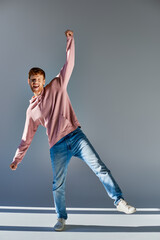 smiling red haired man in casual outfit standing on one leg with one hand raised looking at camera