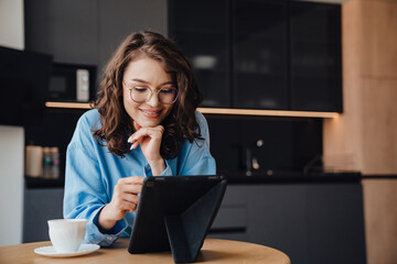 Smiling woman drinking coffee and using tablet computer while sitting in kitchen