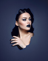 Close-up portrait of a young woman with gothic dark makeup. The face and hands of a woman in a hole made of dark paper. Evening, dark makeup. Advertising of a beauty salon, decorative cosmetics.