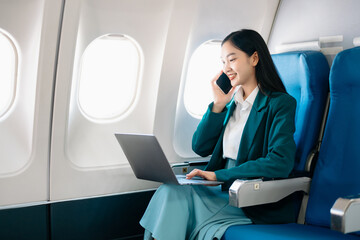 Portrait of  successful Asian businesswoman or entrepreneur in a formal suit on an airplane sitting in business class using a phone, computer laptop. Travel in style, work with grace.