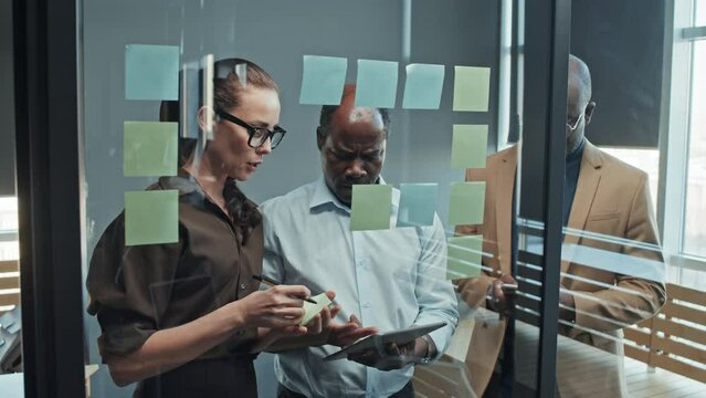 Medium through-glass slowmo shot of multiracial business team. Caucasian female employee gluing stickers on window and black male coworkers using tablet and phone