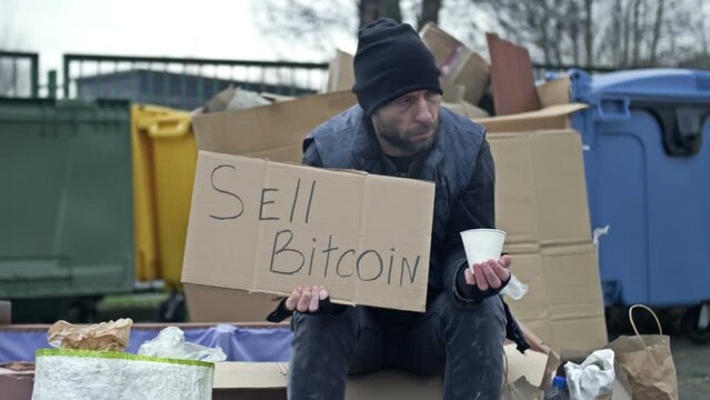 Cold, unkempt-looking young man sits with a begging cup by a pile of rubbish and holds up a handwritten SELL BITCOIN poster.