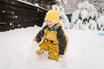 Fototapeta na wymiar Child catching snowflakes tongue in winter clothes having fun. Kid wearing warm hat walking in winter park. Boy fall into snow in mountain country in snowy forest. Baby playing snow among snowdrifts.