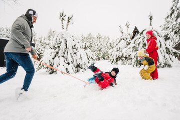 Dad pulling little son on winter day. Children are rolling down hill on sledge in forest. Happy...