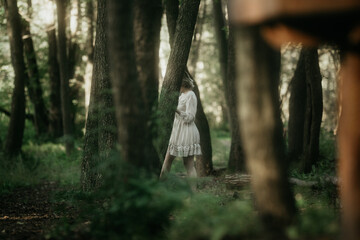 A girl in a white dress walks between the trees in a dark gloomy forest