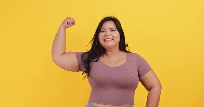 A chubby Asian woman wearing exercise clothes is showing off her physical strength on a yellow background.	