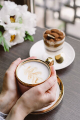 Pink coffee cup on the table in cafe over the window. Flowers and tiramisu served for morning drink. Hands holding cup 