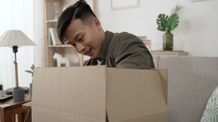 happy Korean male customer sitting down on couch to unpack the parcel carton box is surprised and...