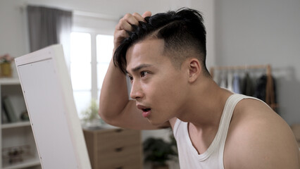 unhappy chinese man checking hairstyle in the mirror is shocked to spot dandruff on his head while preparing for a date in the bedroom at home.