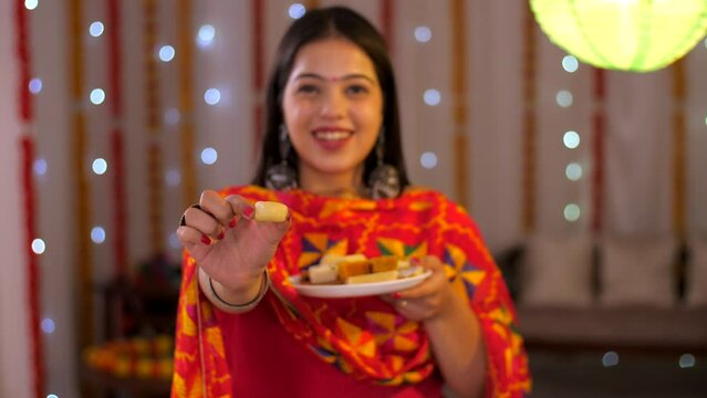 An attractive Indian woman happily smiling and offering sweets - posing for the camera - Diwali festival. Beautiful young female offering barfi with a wide smile on her face - decorative festive ba...