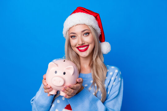 Photo portrait of attractive blonde young woman hold piggy financier wear sweater x-mas hat isolated on blue color background