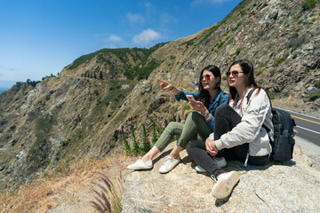 two cheerful asian Chinese women on California road trip sitting on rock enjoying breathtaking landscape along pacific coast highway in western states