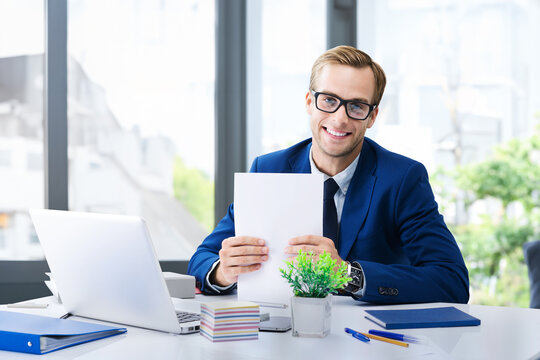 Portrait photo of smiling businessman in glasses, suit work with documents and laptop computer at office workplace. Business, job, banking concept. Executive employee sit against window on background.