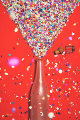 Confetti popping champagne bottle on red background under confetti rain.