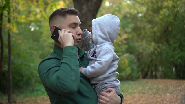 A young father holds his son in his arms and talks on the phone. A man with a child in the park with a smartphone.