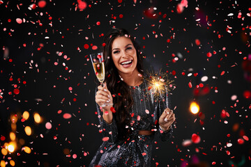A beautiful woman surrounded by confetti, holding a sprinkle and a glass of champagne. New Year's party.