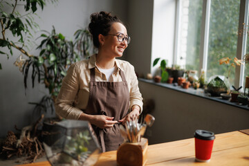 Smiling young asian florist tying apron near coffee to go, gardening tools and plants in floral shop