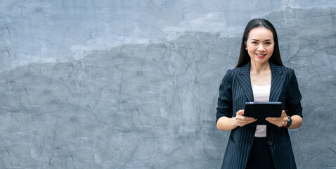 Portrait Asian businesswoman professional in black business suit using a digital tablet standing in...