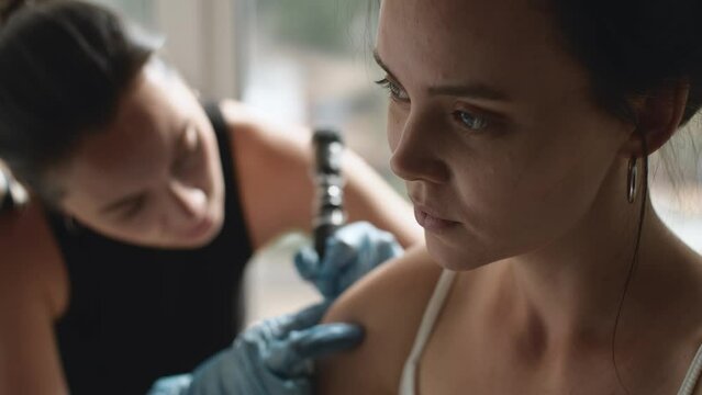 Portrait of beautiful woman while applying tattoo by tattoo artist in a salon. Professional tattooist makes tattooing on the shoulder. Cinematic shot