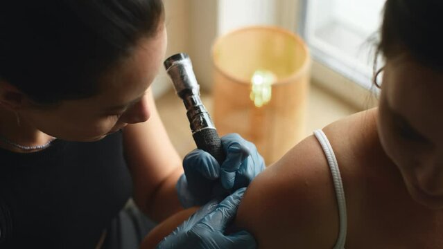 Young woman in tattoo salon during procedure. Professional female tattooist makes tattooing on customers shoulder in tattoo studio. Cinematic shot