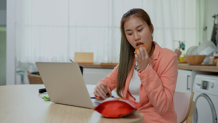 asian young business woman using laptop working and eating snack in Kitchen at home . Asia female...