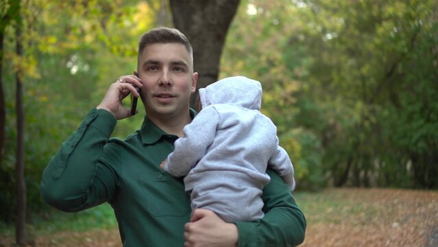 A young father holds his son in his arms and talks on the phone. A man with a child in the park with a smartphone.