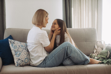 Obraz premium Smiling mommy sitting on sofa with little daughter, showing love. Hilarious mother nanny babysitter tickling small kid girl, having fun together on comfortable couch, enjoying priceless time at home.