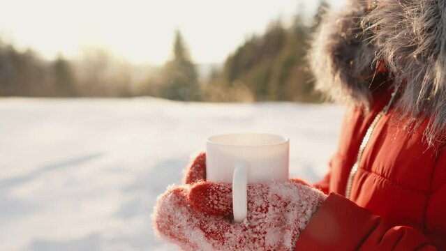 A woman is holding a white cup with a warming hot drink that is steaming in the yard in winter. Delicious tea or aromatist coffee. Mulled wine in the snow is very tasty. Street snack at a picnic