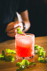 Close up of a barman final touching red icy cocktail with a final mint decoration with scattered mint leaves on the wooden counter and bartender hand in the back