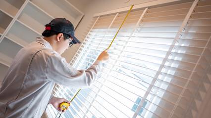 Home renovation or House remodeling concept. Asian male furniture assembler or Interior construction worker man using tape measure on metal white blinds on the window.