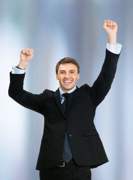 Photo of very happy, excited gesturing young business man in black confident suit, raising hand fist, against blurred office background. Advertisement ad concept.