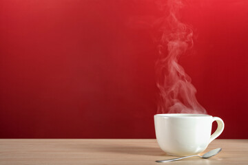 Steam rising from a large white porcelain cup of hot coffee or tea sitting with a silver spoon on a blonde colored wooden table against a red wall