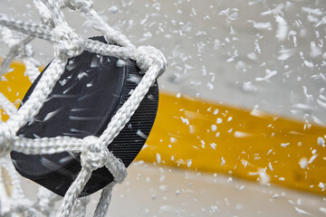 A close-up view of an Ice Hockey puck hitting the back of the goal net as shavings fly by, viewed from the side. Scoring a goal in ice hockey.